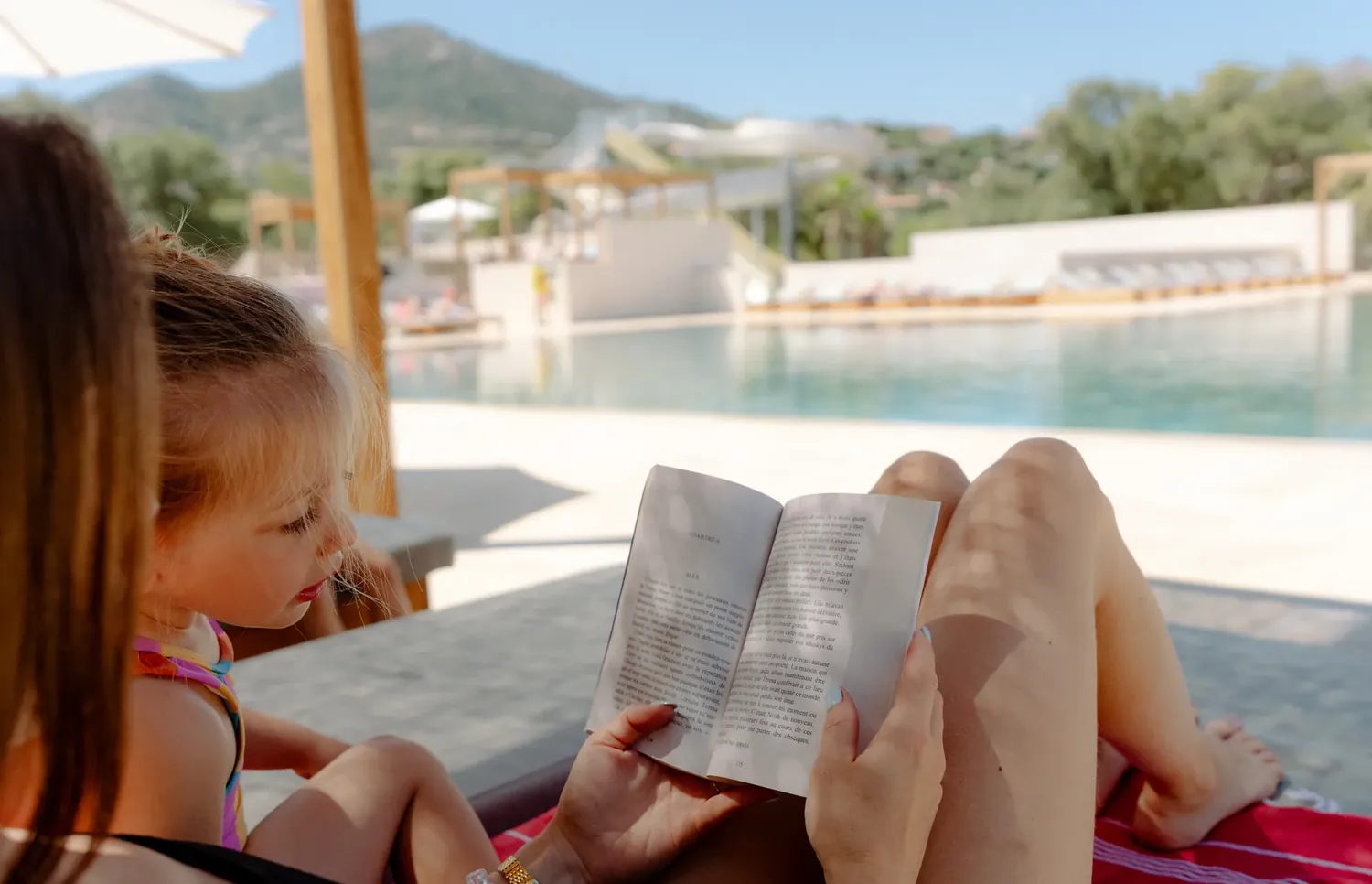 Une mère et sa fille lisant un livre ensemble sur un transat au bord de la piscine du camping en Balagne.